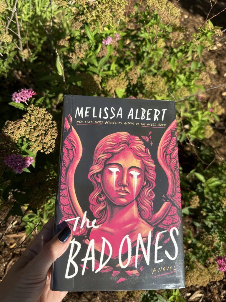 The book the Bad Ones by Melissa Albert held over some pink flowers. The cover shows a pink statue of a crying angel