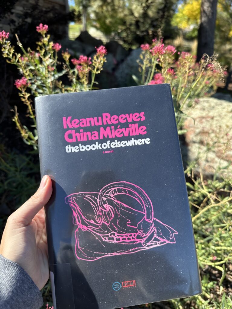 The Book of Elsewhere held up in front of pink flowers. The cover is black with a sketch of a babirusa skull in pink