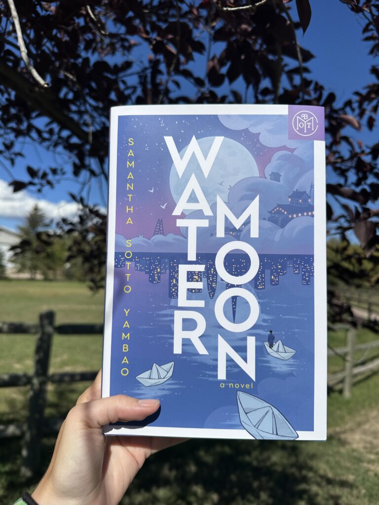 The book Water Moon by Samantha Sotto Yambao held outside. The cover shows a dreamy cloudscape and the reflection of a city with paper boats floating in the water
