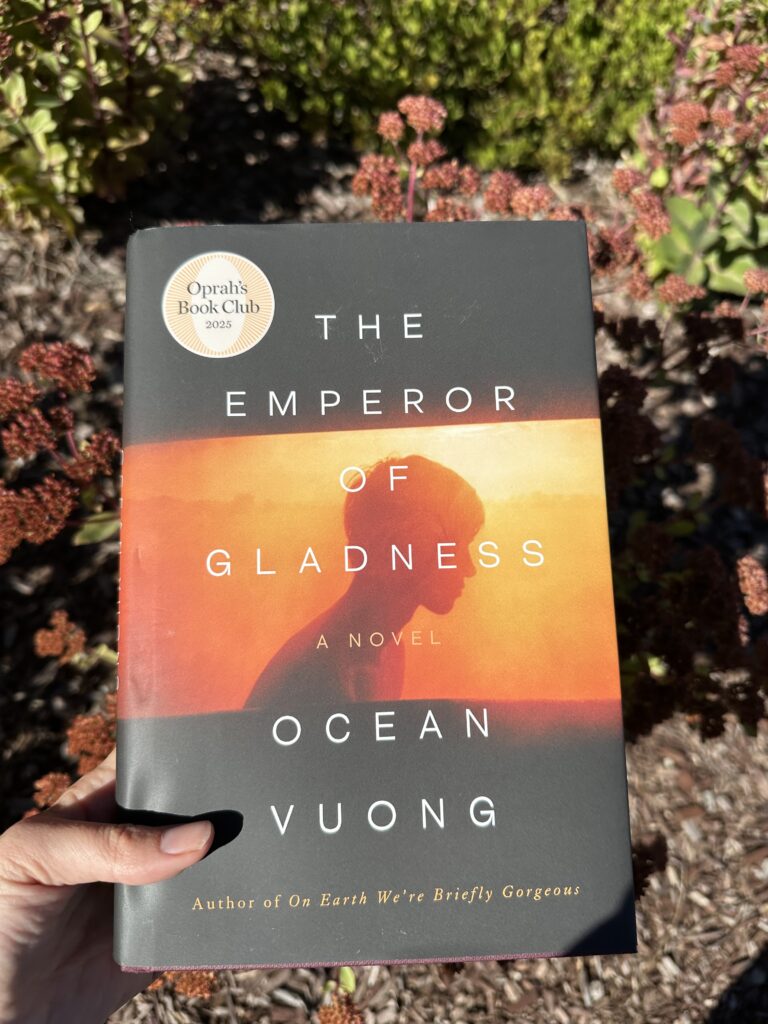 The book The Emperor of Gladness by Ocean Vuong held over some dying flowers. The cover shows a young man's silhouette against a setting sun