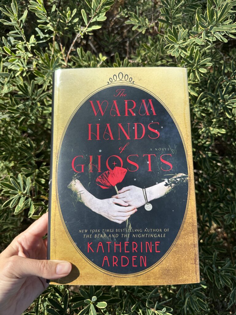 Held over a green bush, the book The Warm Hands of Ghosts by Katherine Arden shows two ghostly hands emerging from darkness holding a poppy flower