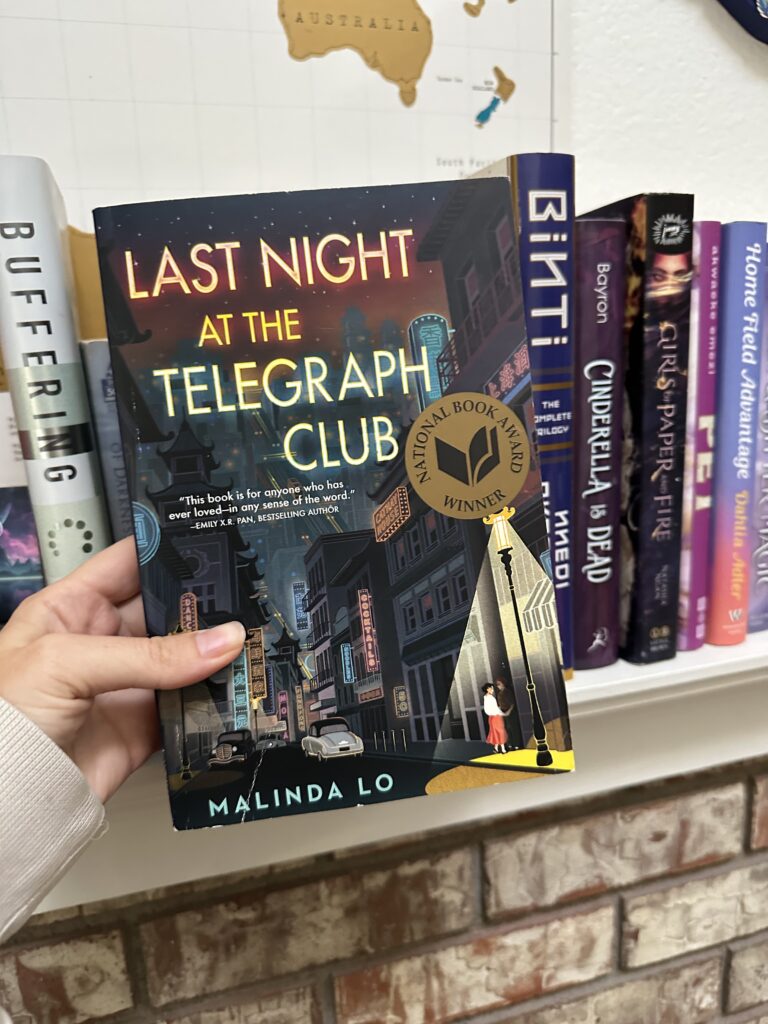 The cover of Last Night at the Telegraph Club by Malinda Lo shows a neon-lit Chinatown street at light with a couple under a streetlight