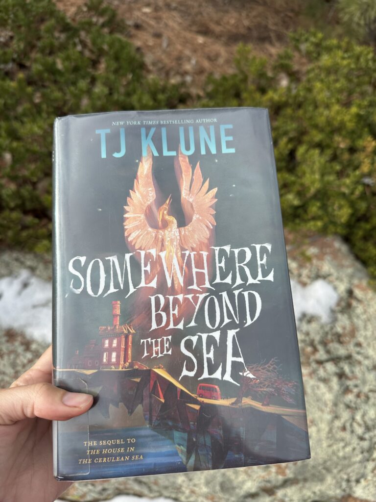 The cover of Somewhere Beyond the Sea by TJ Klune shows a house out on a promontory of rock with a phoenix rising above it and a red bus driving away from the house
