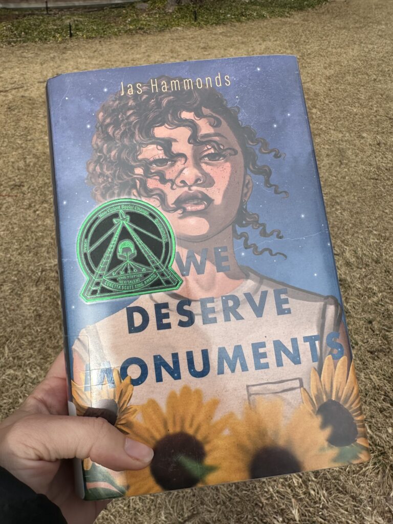 The cover of We Deserve Monuments by Jas Hammonds shows a light brown skinned girl with freckles and curls looking out at the future over a background of stars and a foreground of sunflowers