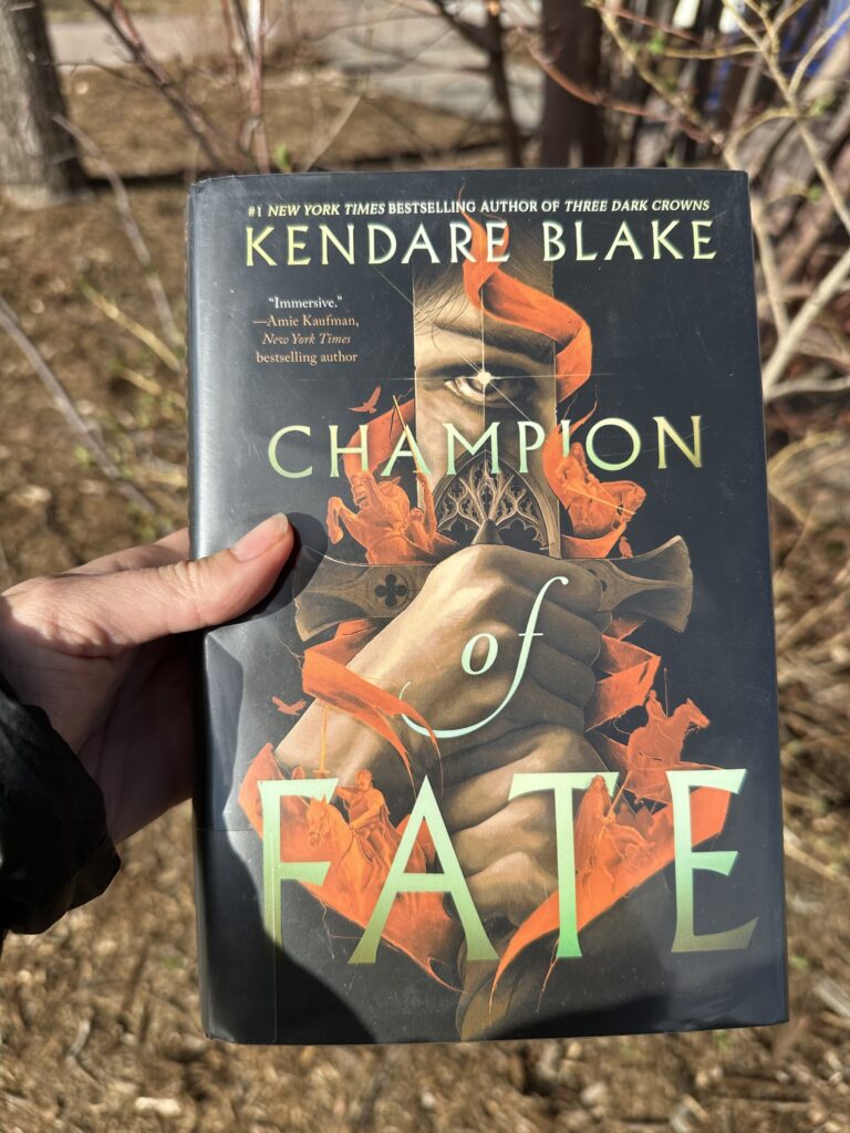 The cover of Champion of Fate by Kendare Blake shows two hands holding the hilt of a sword with an eye reflected in the sword and shadowy figures on horseback twisting around the hands and blade