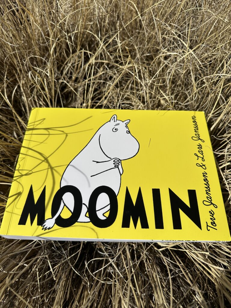 The cover of Moomin by Tove and Lars Jansson is yellow and shows the titular character Moomin, a round, lovable, hippo-like creature