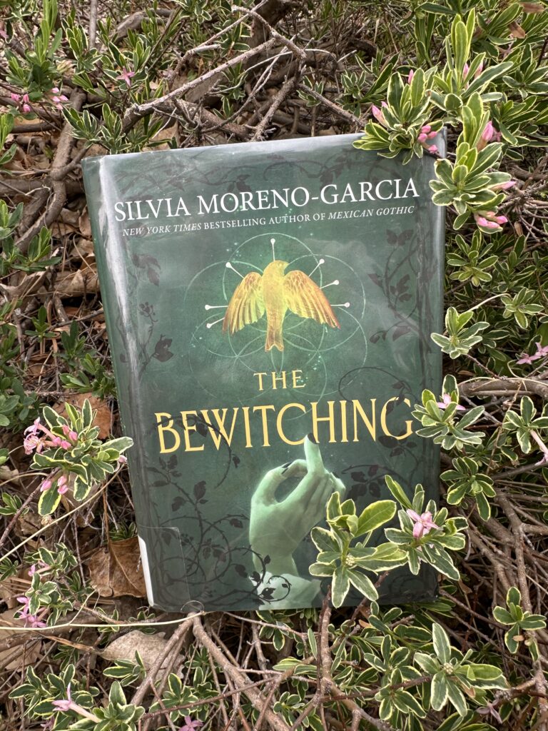 The cover of The Bewitching by Silvia Moreno Garcia shows a ghostly green hand with black nails reaching toward a bird stuck with pins