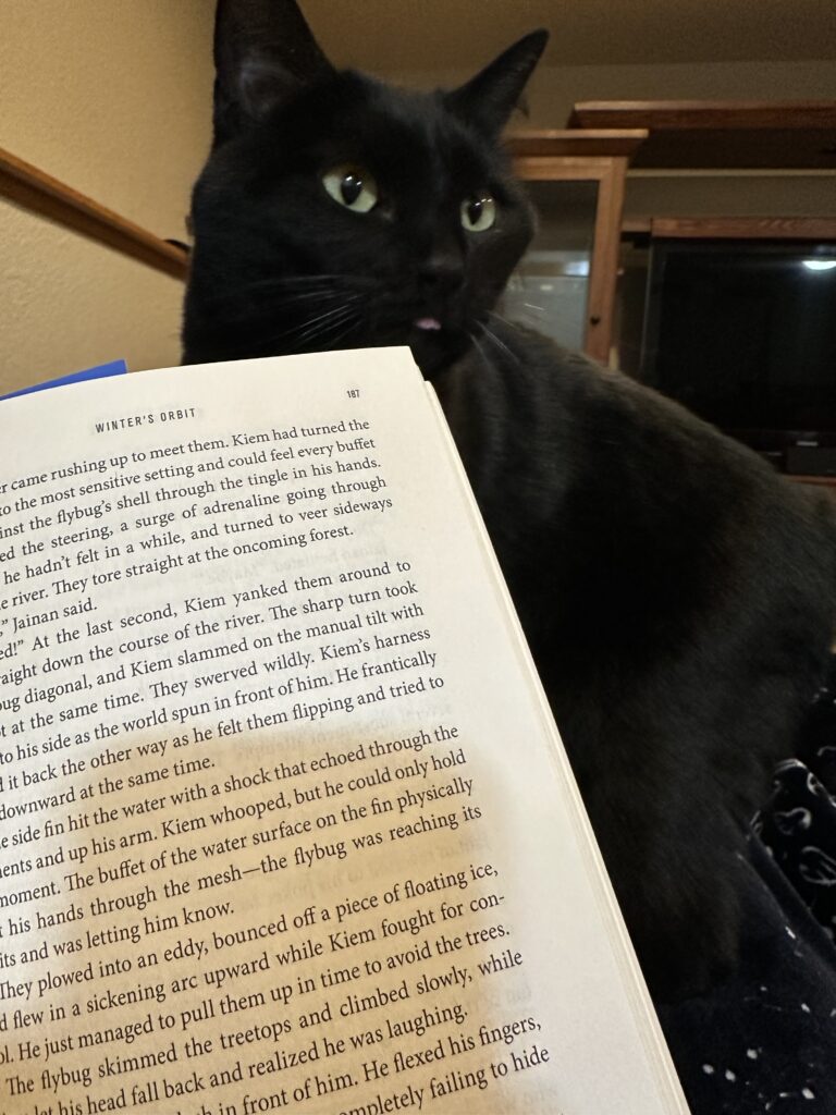 A black cat with its tongue just poking out peers over the corner of an open copy of Winter's Orbit