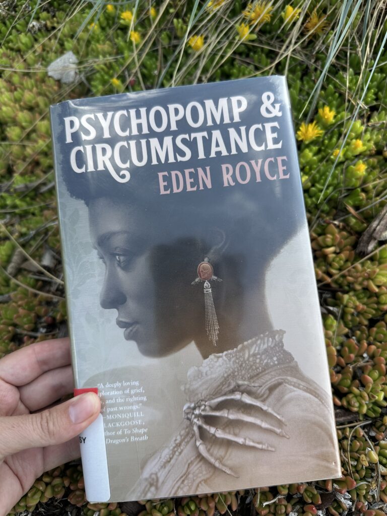 The cover of Psychopomp and Circumstance shows the profile of a young Black woman dressed in early 20th century fashion. Her earrings have a skull on them and her broach is the shape of a skeleton hand.