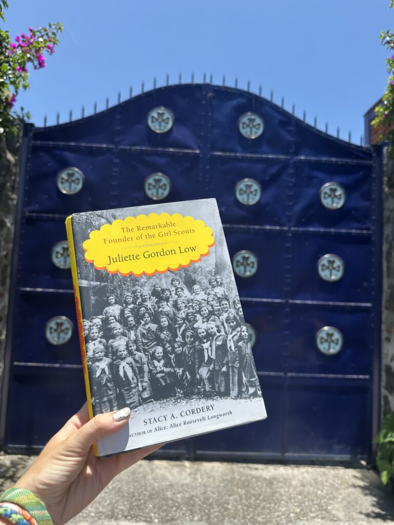 A hand holds up a copy of Juliette Low’s biography before a set of blue double doors covered with trefoil emblems of Girl Scouts