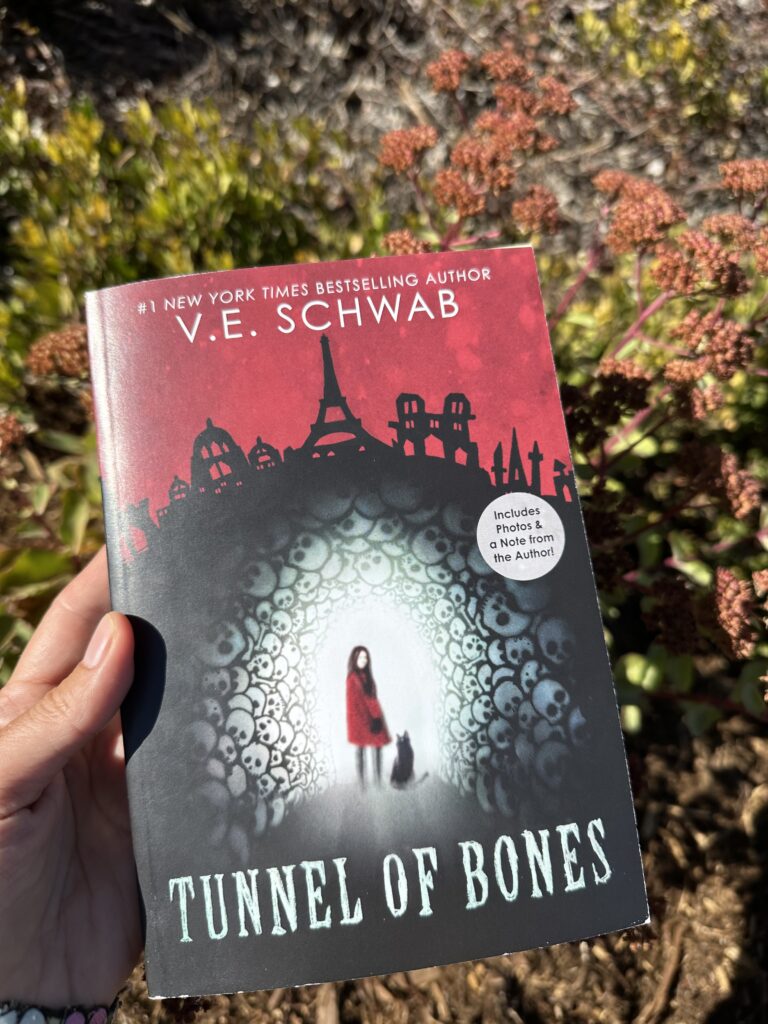 On the cover of Tunnel of Bones, a girl and her cat stand in a tunnel made of skulls beneath the Paris skyline